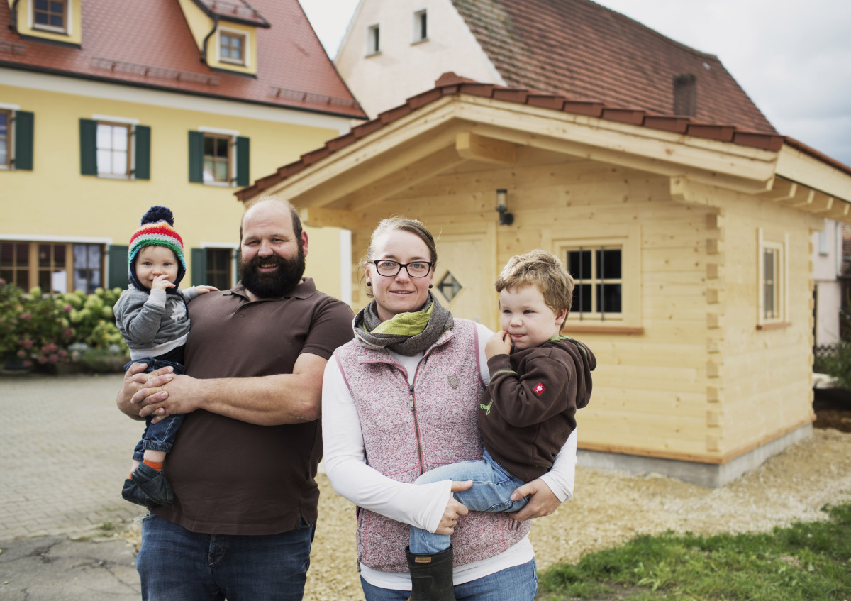 Zwei erwachsene Personen stehen mit zwei Kindern, die sie jeweils im Arm halten, vor einem Häuschen aus Holz.