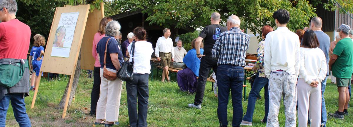 Personen stehen auf der Wiese und blixken auf einen Mann, der spricht.