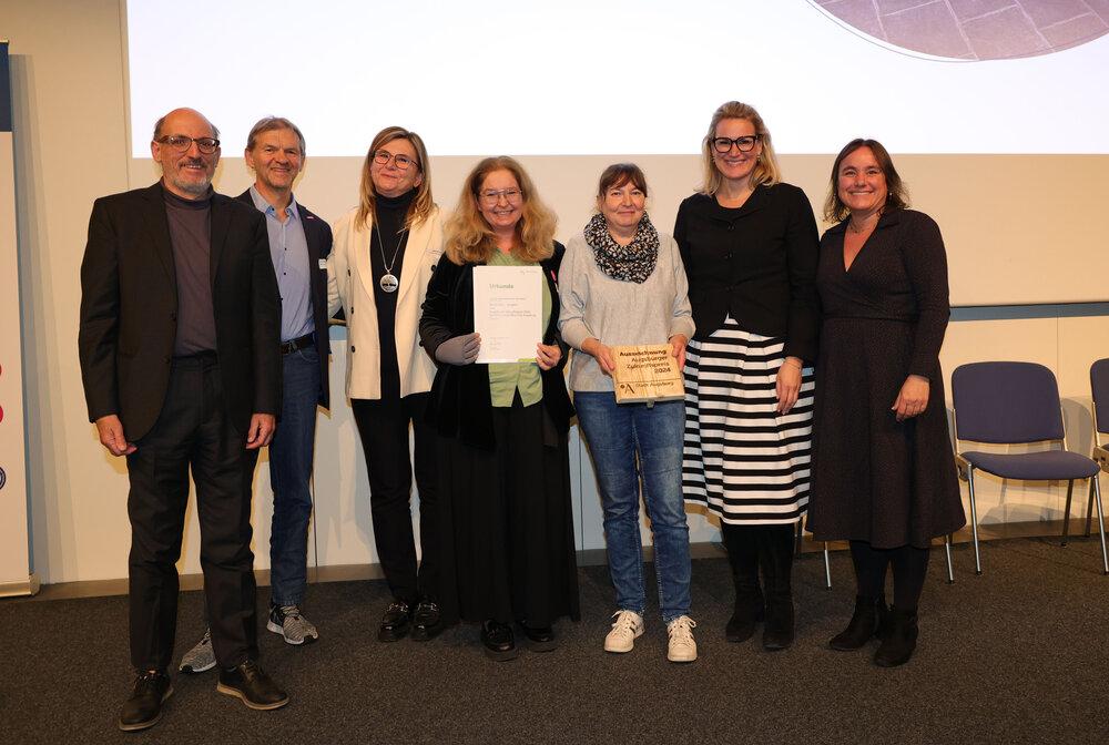 Das Foto zeigt mehrere Personen auf einer Bühne. Eine Person hält eine Urkunde in der Hand.