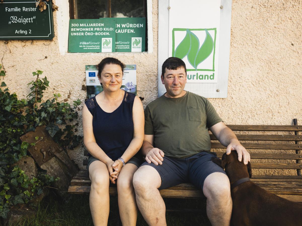 Eine Frau und ein Mann sitzen auf einer Bank von einer Hauswand. Der Mann hat seine linke Hand auf den Kopf eines Hundes gelegt.