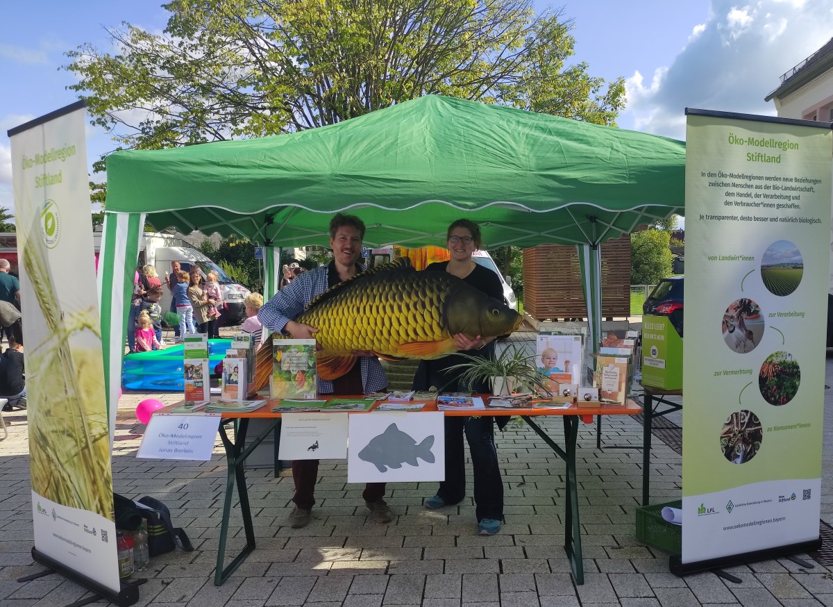 Projektmanager*in posieren hinter dem Stand der ÖMR Stiftland mit großem Stoff-Karpfen