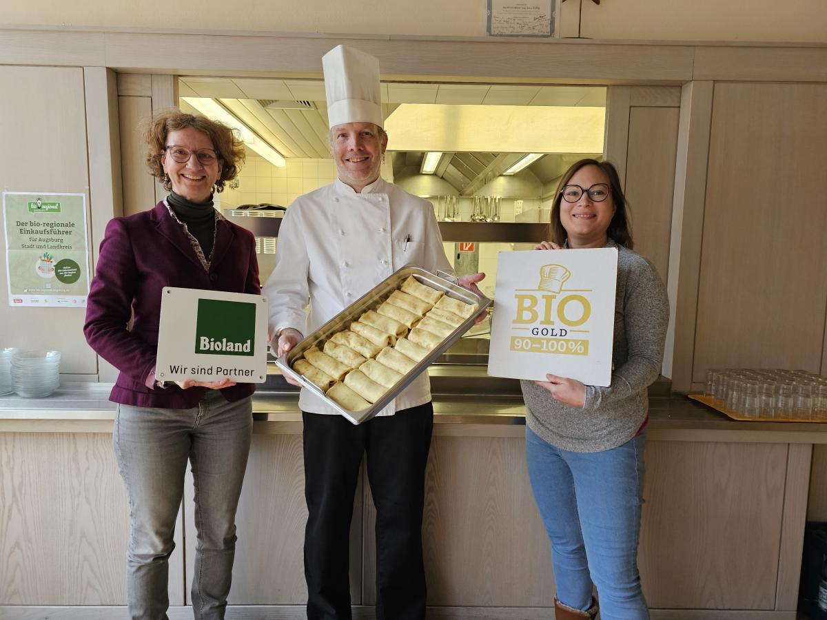 Das Foto zeigt einen Koch, der Essen in der Hand hält und zwei Frauen die Schilder hochhalten.