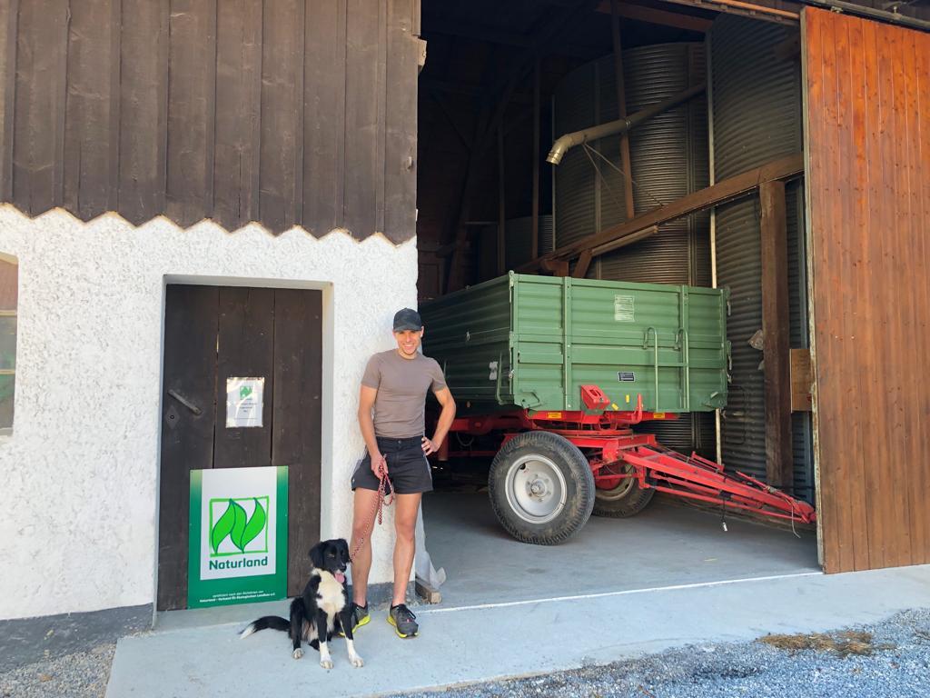 Das Foto zeigt einen Mann mit Hund vor einem Landwirtshcaftlichen Gebäude