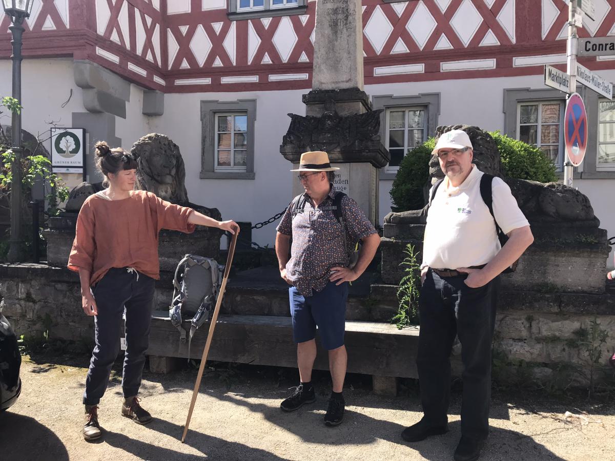 Eine Frau mit einem Stab in der Hand, daneben zwei Männer, im Hintergrund ein Brunnen und Fachwerkhäuser