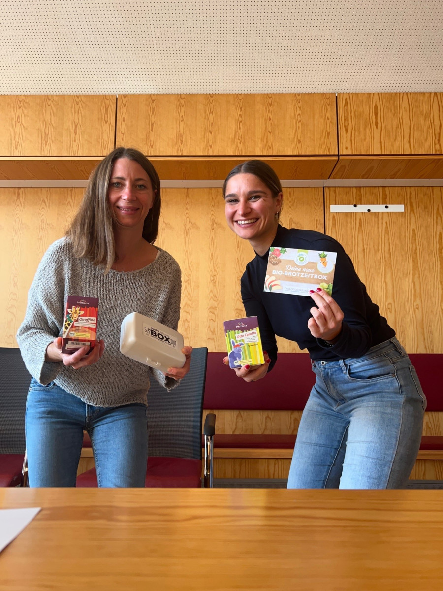 Zwei Frauen halten eine Teepackung, eine Brotzeitbox und einen Flyer in der Hand