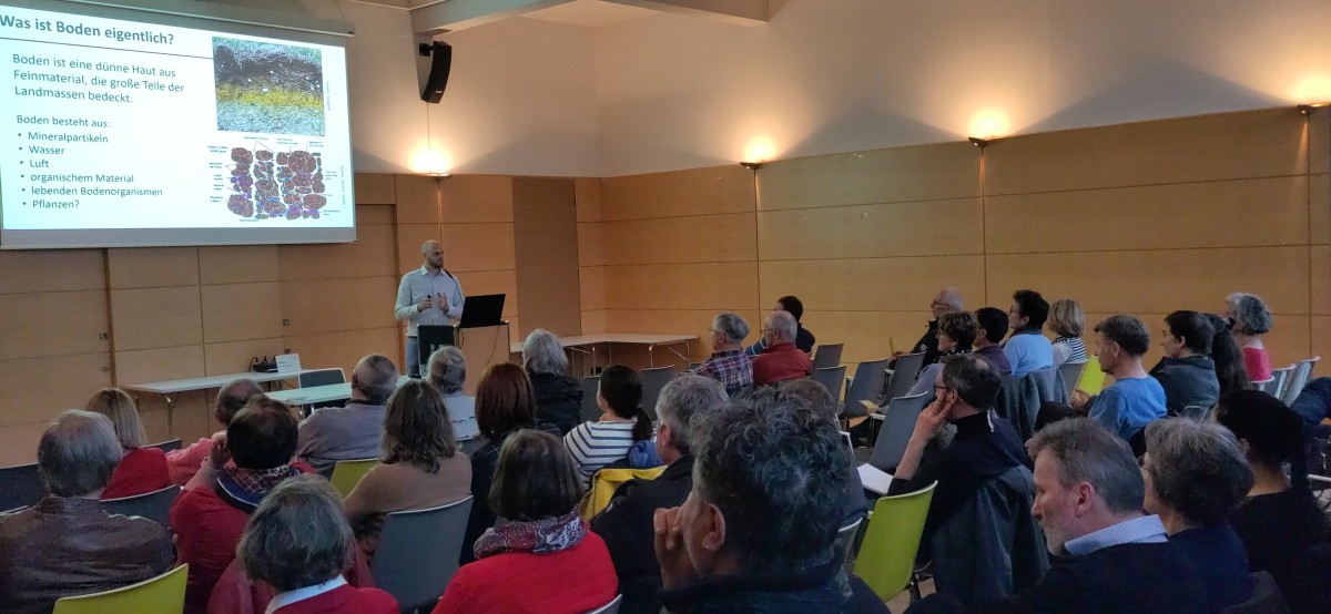 Blick in den Saal mit Publikum, Dr Löbmann und einem Ausschnitt seiner Präsentation