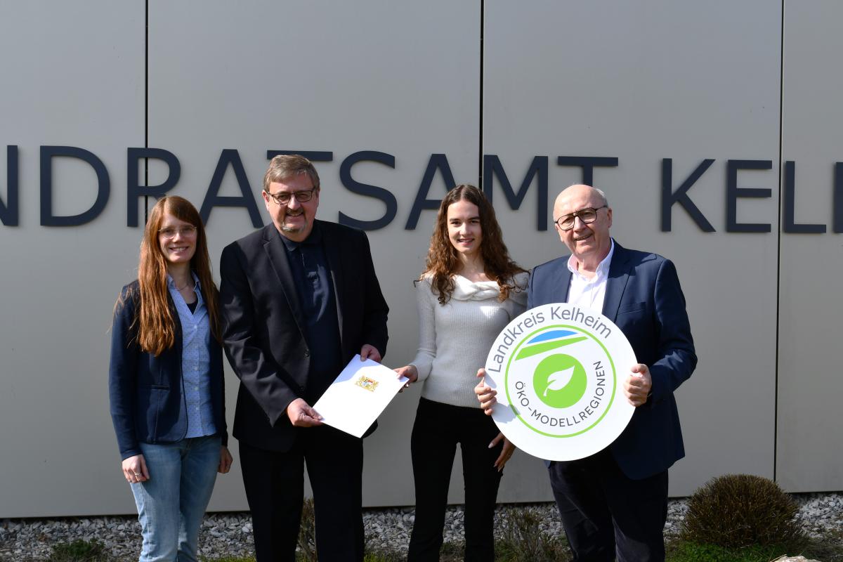 Vier Personen vor dem Landratsamt Kelheim, einer übergibt symbolisch die Urkunde und einer hält das Schild mit dem Logo der Öko-Modellregionen