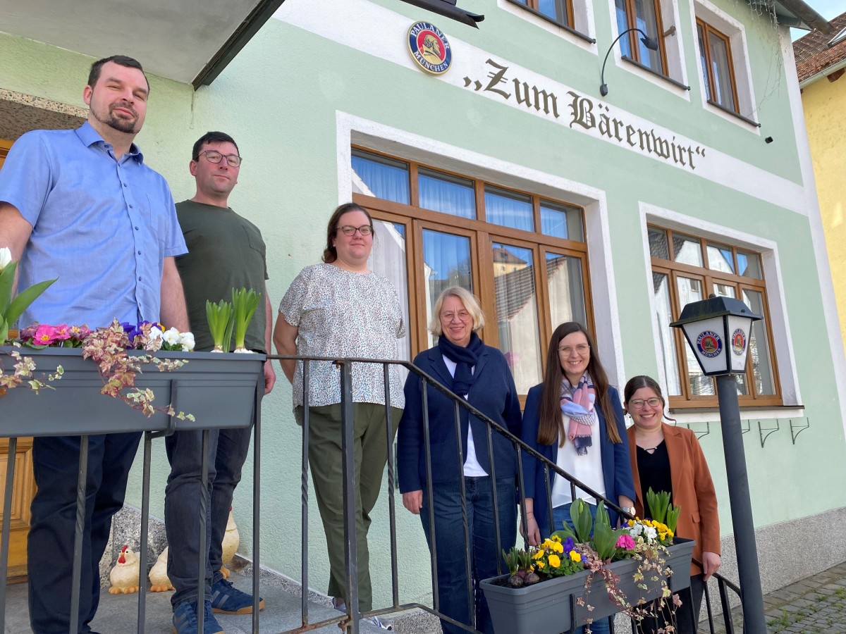 Mehrere Personen stehen vor einem Wirtshaus auf einer Treppe.