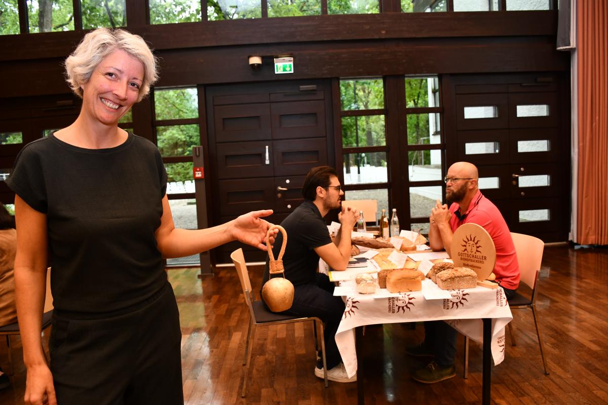 Frau mit Kuhglocke in der Hand, im Hintergrund Tisch mit Lebensmitteln und 2 Männern