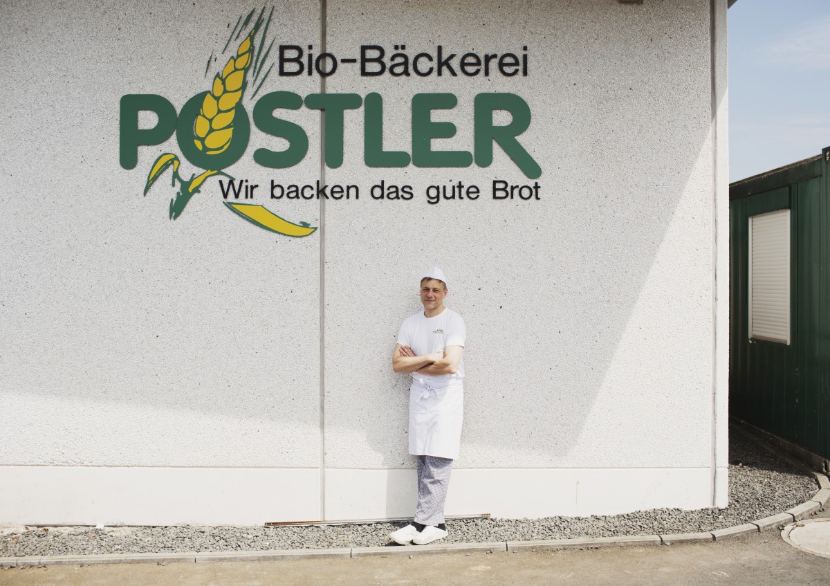 Roland Postler steht vor der Backstube in Burgebrach, an der Hauswand sieht man den Schriftzug Bio-Bäckerei Postler