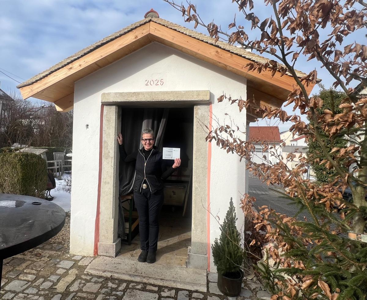 Andrea Beck steht mit der Förderplakette in der Hand in der Tür des Backhäusels