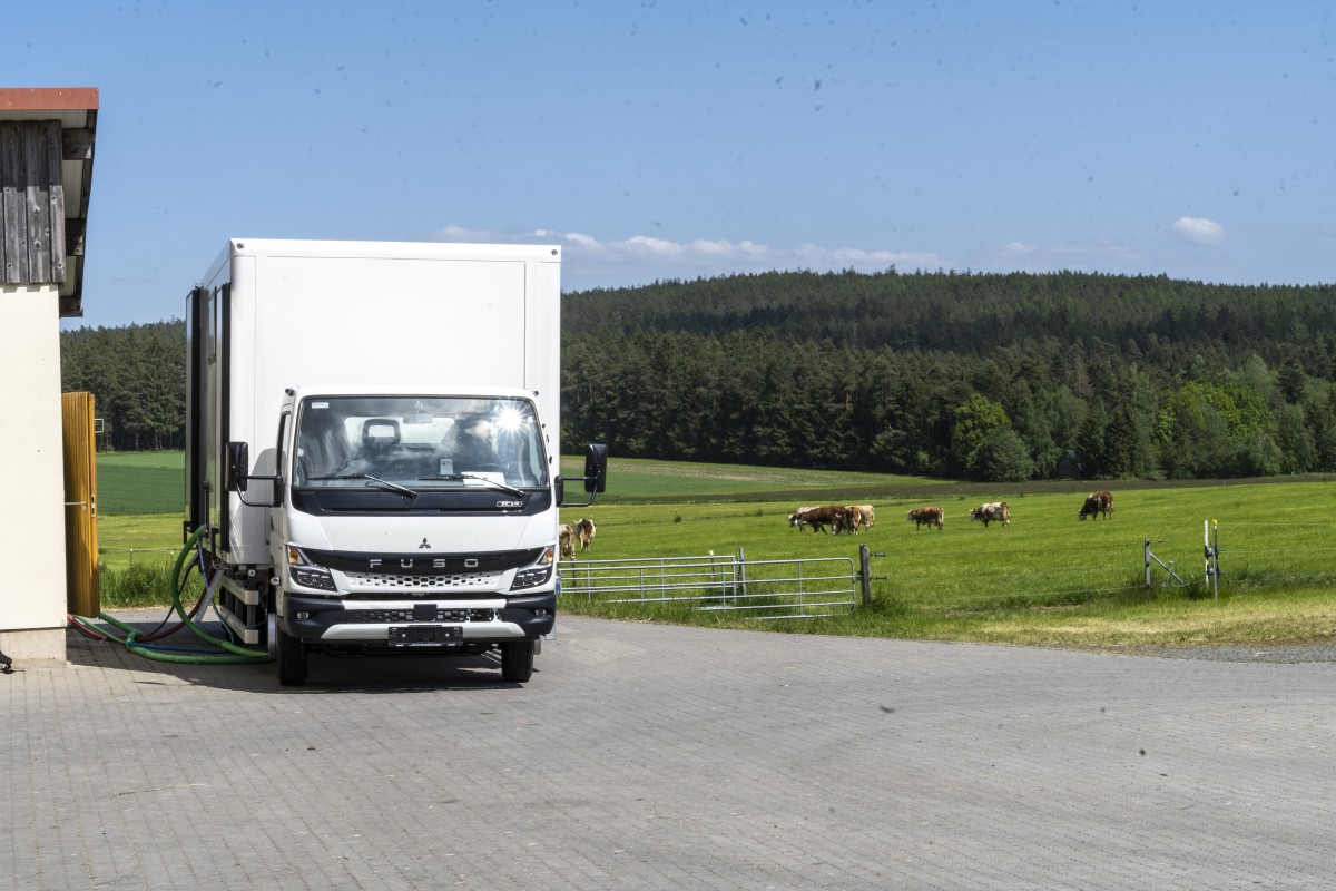 ein weißer LKW steht auf einen landwirtschaftlichen Betrieb, im Hintergrund stehen Kühe auf der Wiese und weiden