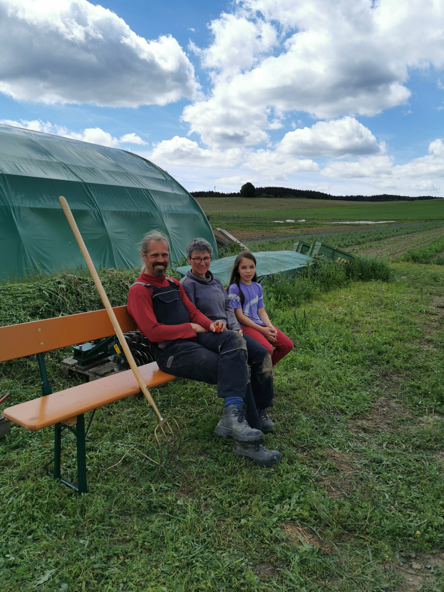 Das Foto zeigt zwei Erwachsene und ein Kind, die auf einer Bank auf einem Feld sitzen.