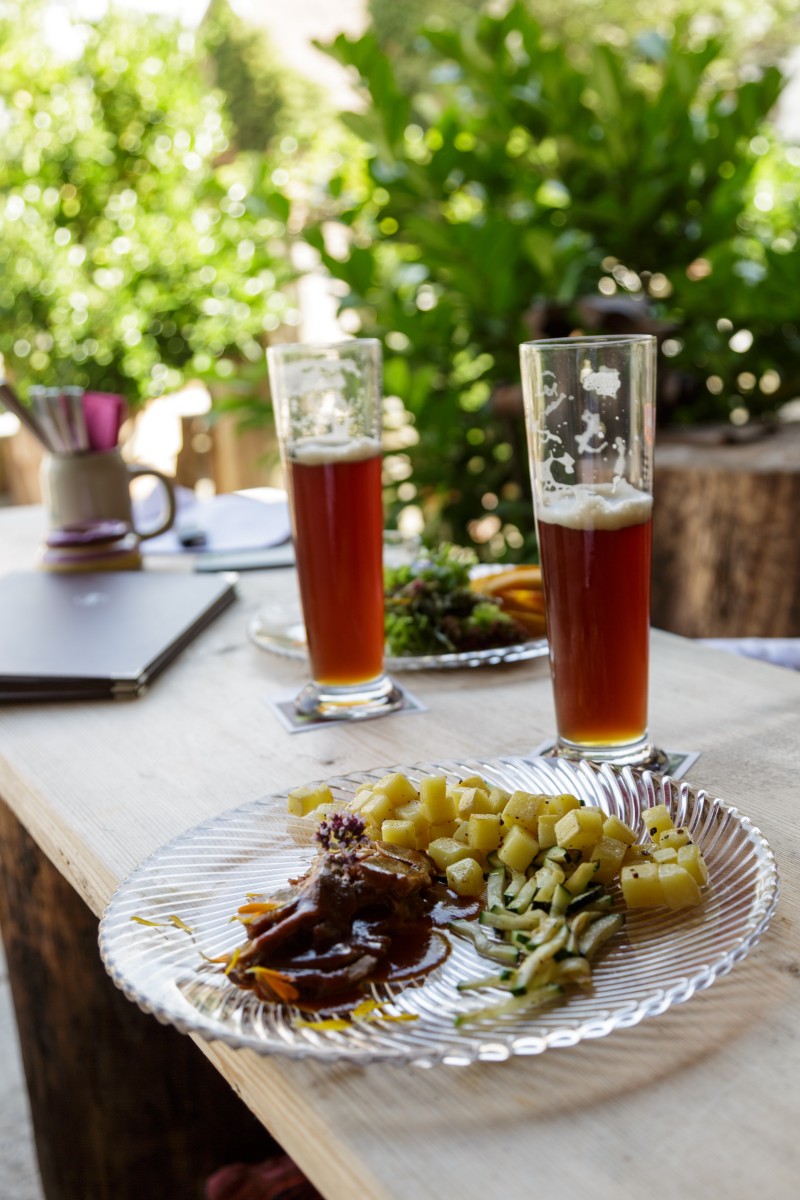 Ein gefüllter Teller steht auf einem Holztisch, im Hintergrund ist ein zweiter Teller und zwei Bier zu sehen.