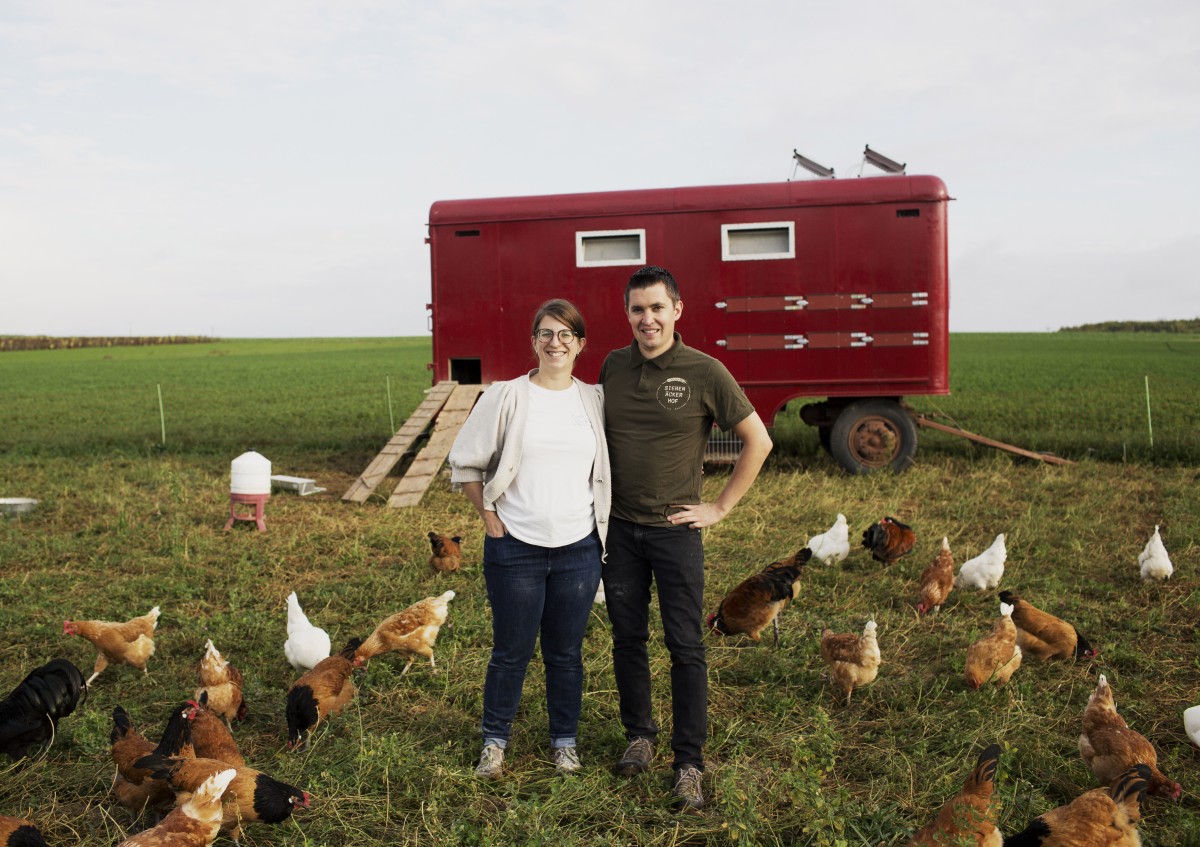 Zwei Menschen auf der Wiese, im Hintergrund ein mobiler Hühnerstall und Hühner auf der Wiese