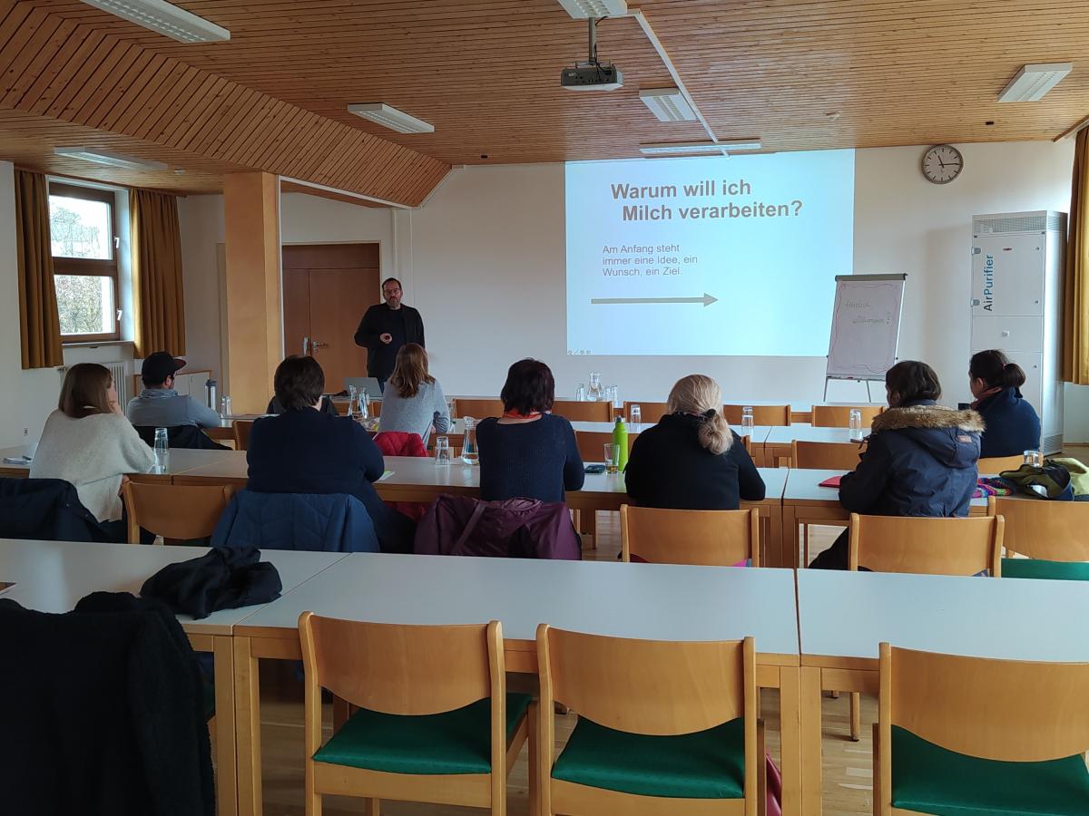 Mehrere Menschen sitzen hinter Tischen und hören einem Referenten zu, der vor einer Präsentation steht.