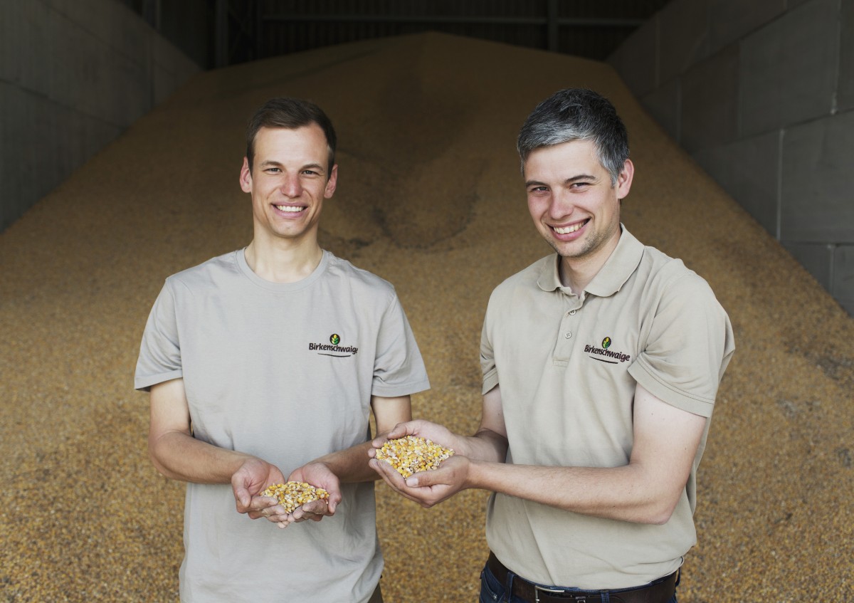 Christian und Stefan Froschmeir halten lächelnd Mais in den Händen, im Hintergrund ist eingelagerter Mais zu sehen
