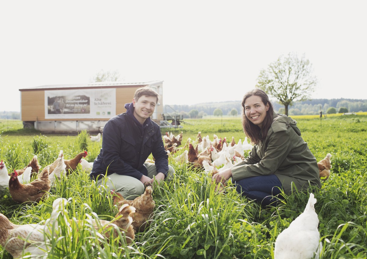 Das Foto zeigt einen Mann und eine Frau auf einer Wiese. Mehrere Hühner umgeben die beiden.