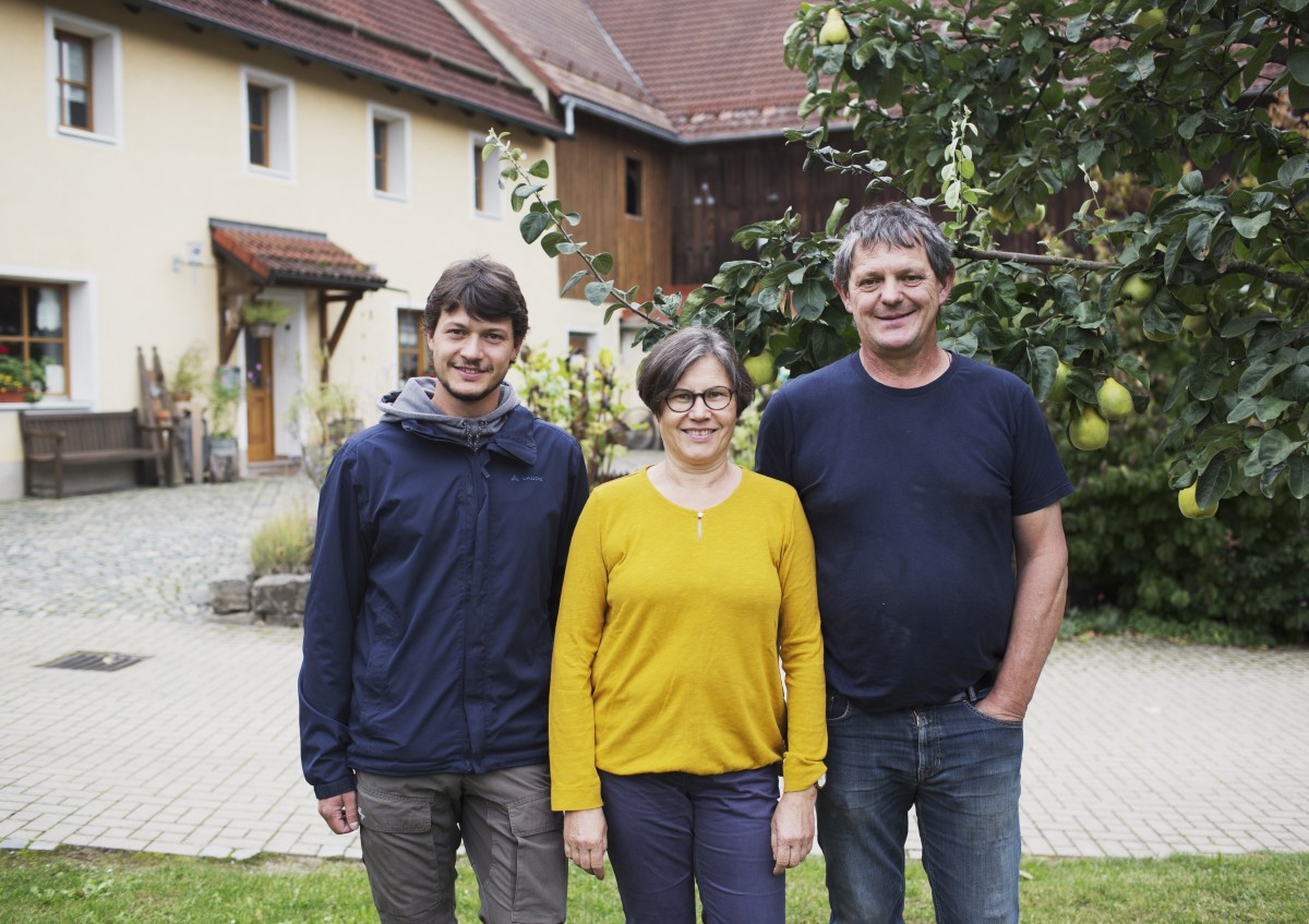 Drei Personen stehen nebeneinander. Im Hintergrund befinden sich mehre Gebäude und ein großer Baum.