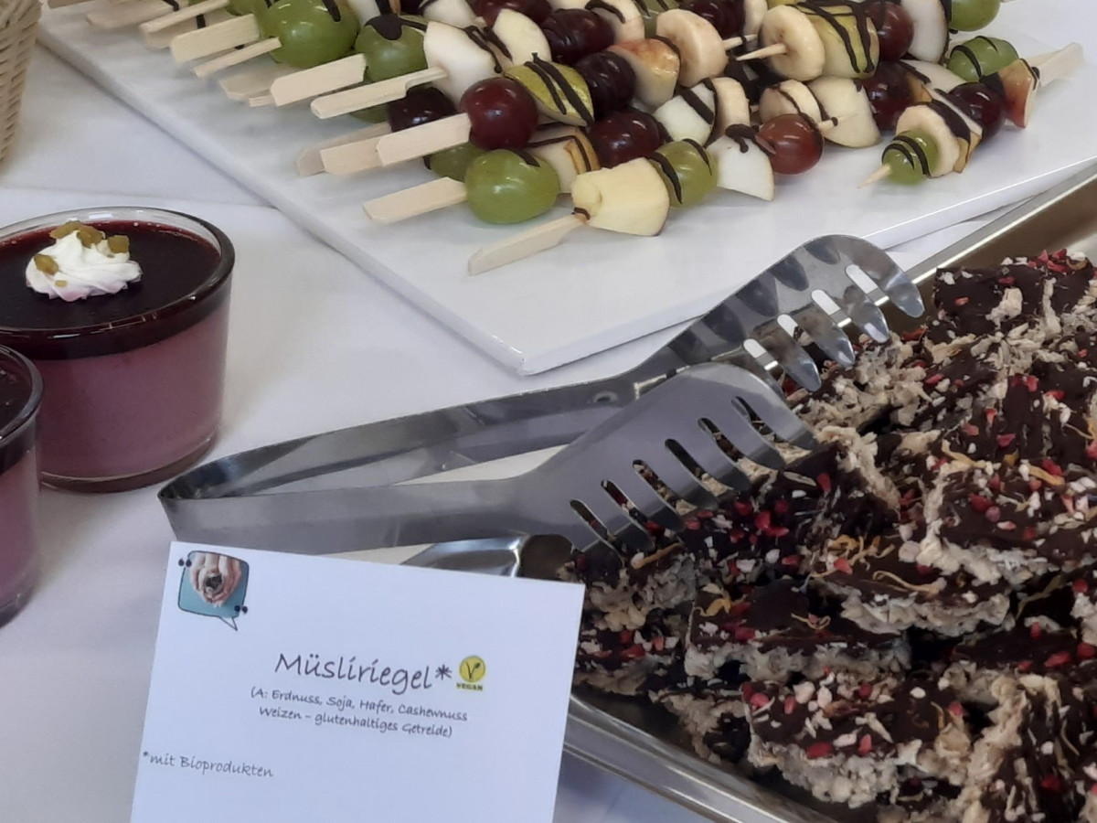 auf dem Bild sieht man Müsliriegel, Kuchenschnitten und Obstspieße sowie ein Himbeerdessert. Ein Schild weißt auf die Verwendung von Bio-Zutaten in Müsliriegel hin