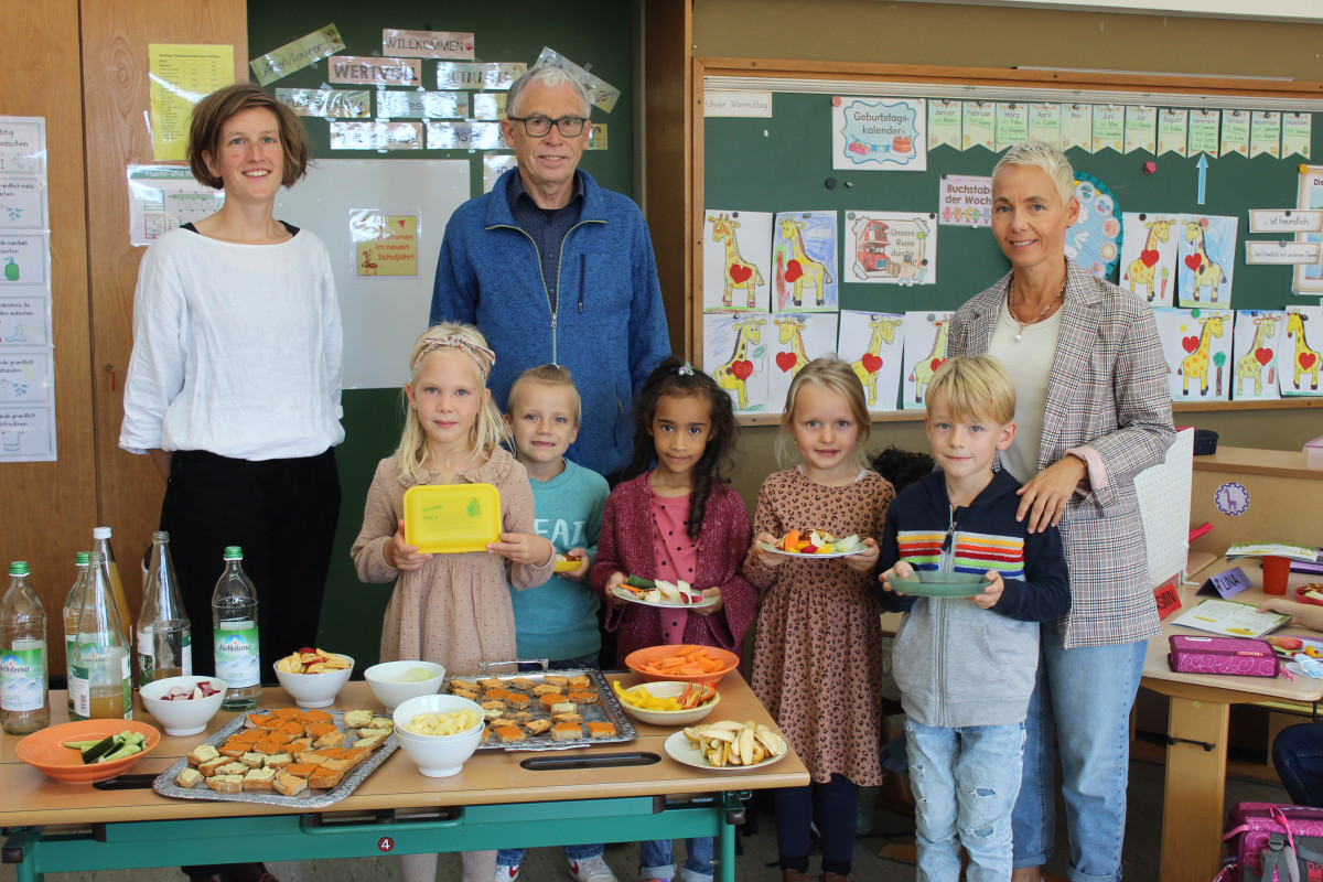 Drei Erwachsene und Kinder hinter einem bunten Büfett mit Brot, Gemüse, Obst