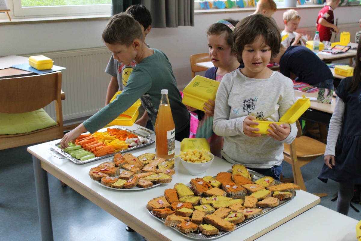 Kinder vor einem Büfett mit einer gelben Box in der Hand: Vor ihnen bunte Häppchen