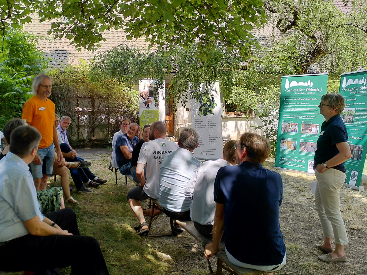 Mehrere Menschen sitzen auf Bänken, diskutierend, im Hintergrund Roll-Ups mit Informationen zum Gut Obbach