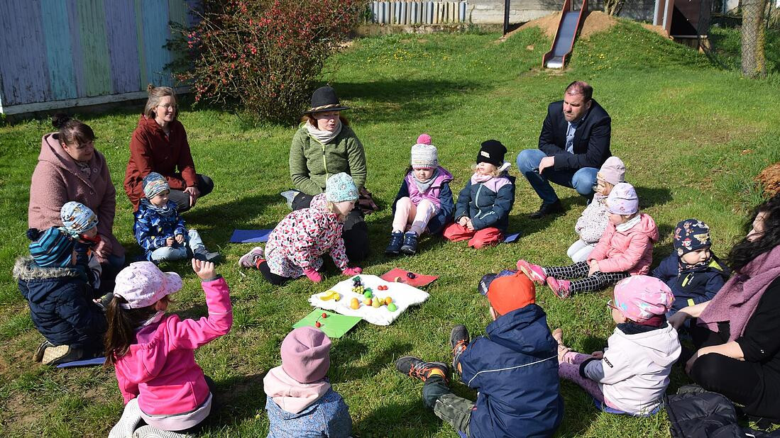 Kinder in einem Sitzkreis auf der Wiese, in der Mitte Holzobst, dahinter Erwachsene