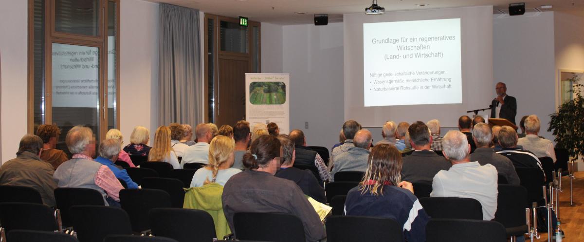 Gäste sitzen im Saal, vorne steht der Referent mit seiner Präsentation.
