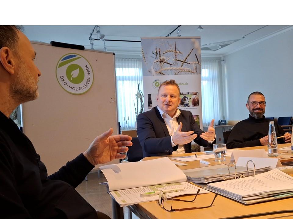 Drei Personen am sitzen am Tisch, auf dem Papiere, Leitz-Ordner und Mineralwasser steht.