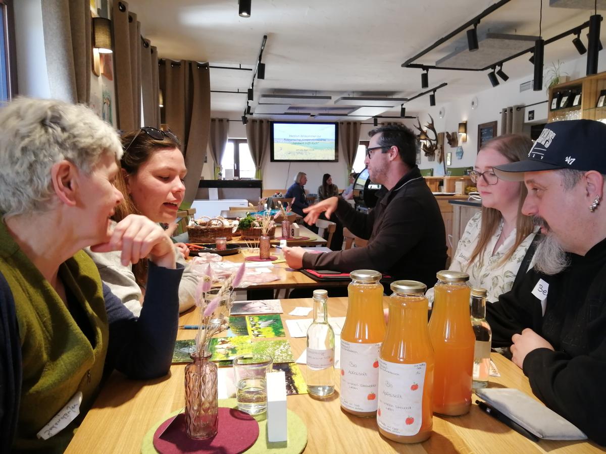 Im Bild links sitzen zwei Frauen, die sich mit einer Frau und einem Mann (rechts im Bild) unterhalten. Auf dem Tisch stehen mehrere Flaschen Streuobstsaft.