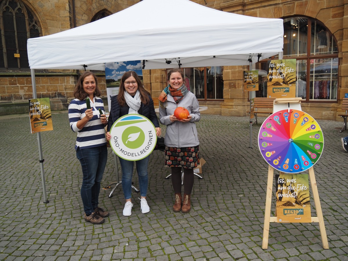 Drei Frauen stehn mit verschiedenen Gegenständen in den Händen vor einem Pavillon. Rechts im Vordergrund ist ein Glücksrad platziert.