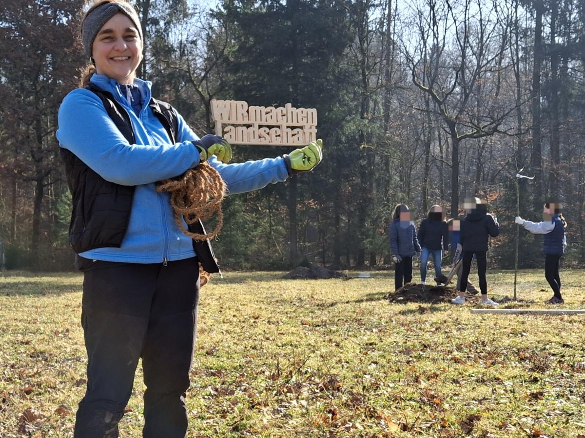 Frau steht auf Wiese mit Holzschilld "Wir machen Landschaft", im Hintergrund sind Kinder, die einen Baum pflanzen.