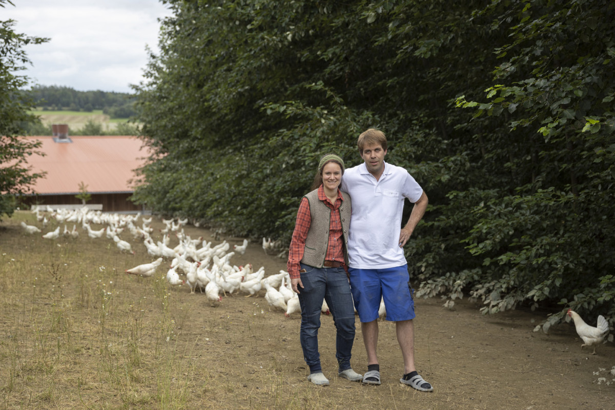 Das Fotos zeigt eine Frau und einen Mann vor Bäumen und Hühnern.