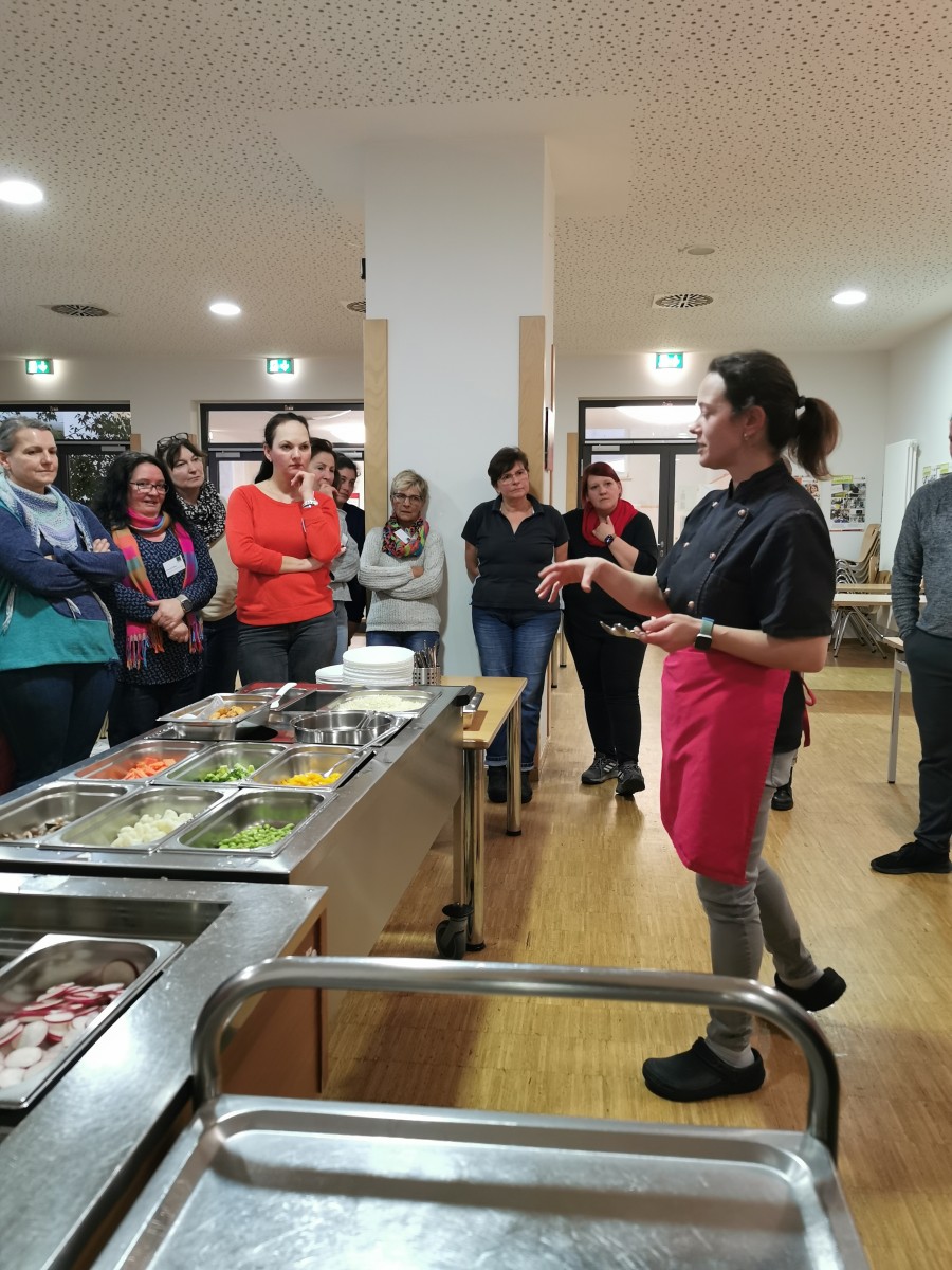 Das Foto zeigt eine Frau, die einer Gruppe von Personen etwas erklärt. Vor ihr ist Essen aufgebaut.